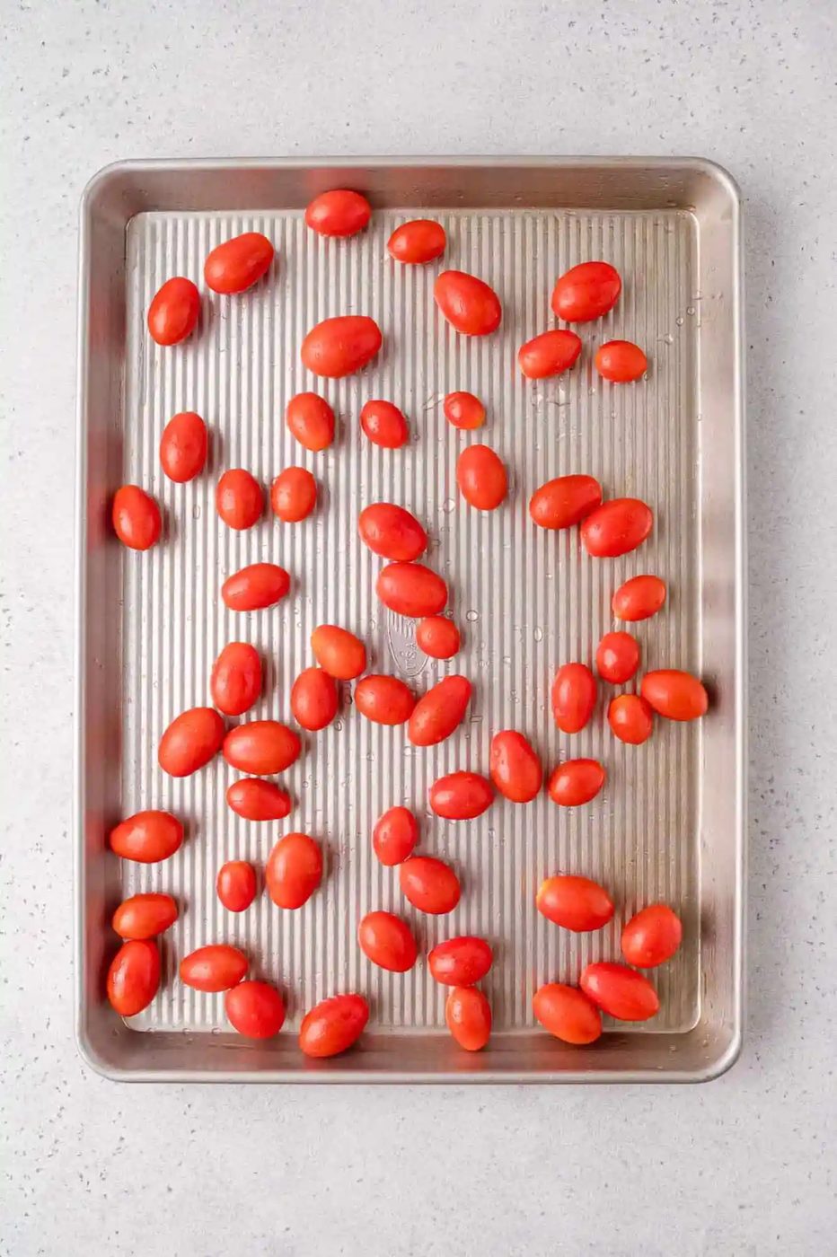 A baking sheet with raw grape tomatoes spread out evenly on a light-colored countertop.