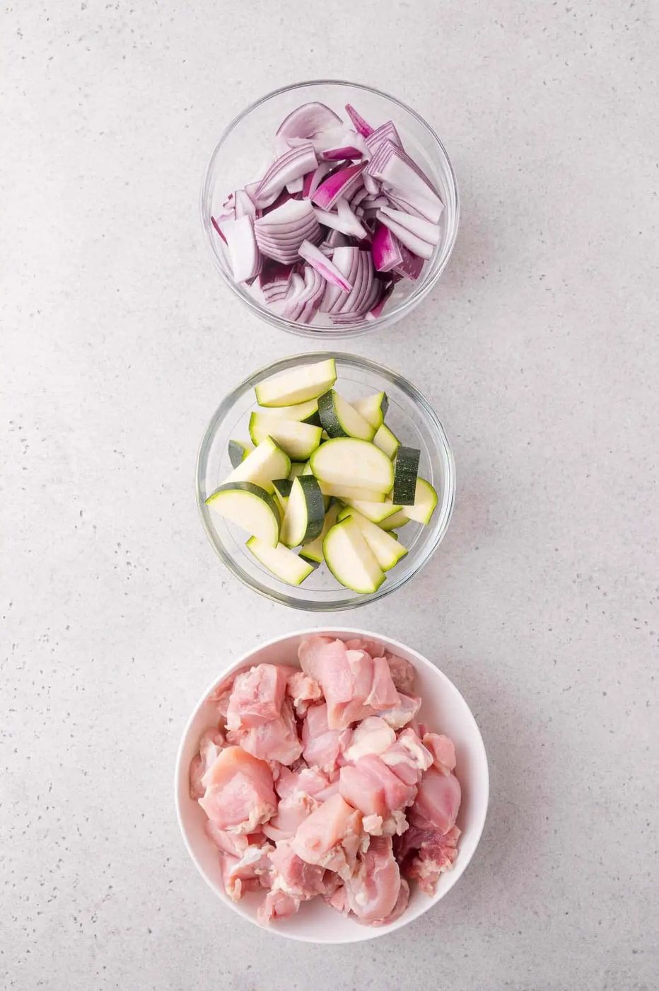 Three bowls containing chopped red onions, sliced zucchini, and raw diced chicken arranged vertically on a light gray surface.