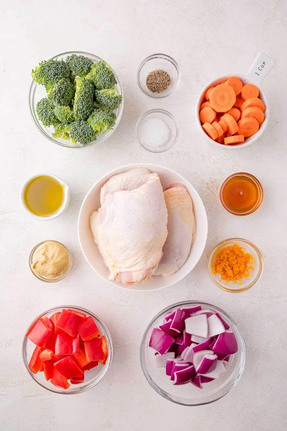 Ingredients arranged in bowls on a white surface, including raw chicken thighs, broccoli, carrots, red bell pepper, red onion, olive oil, mustard, honey, orange zest, salt, and pepper.