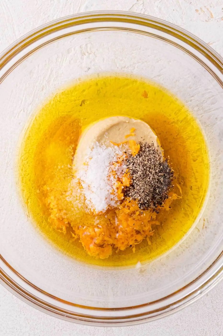 A glass bowl containing olive oil, mustard, salt, black pepper, and citrus zest on a light-colored surface.