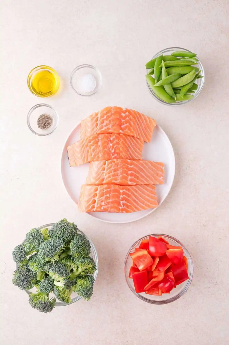A plate of raw salmon fillets on a white surface, surrounded by bowls of broccoli, red bell pepper, snap peas, olive oil, salt, and pepper.