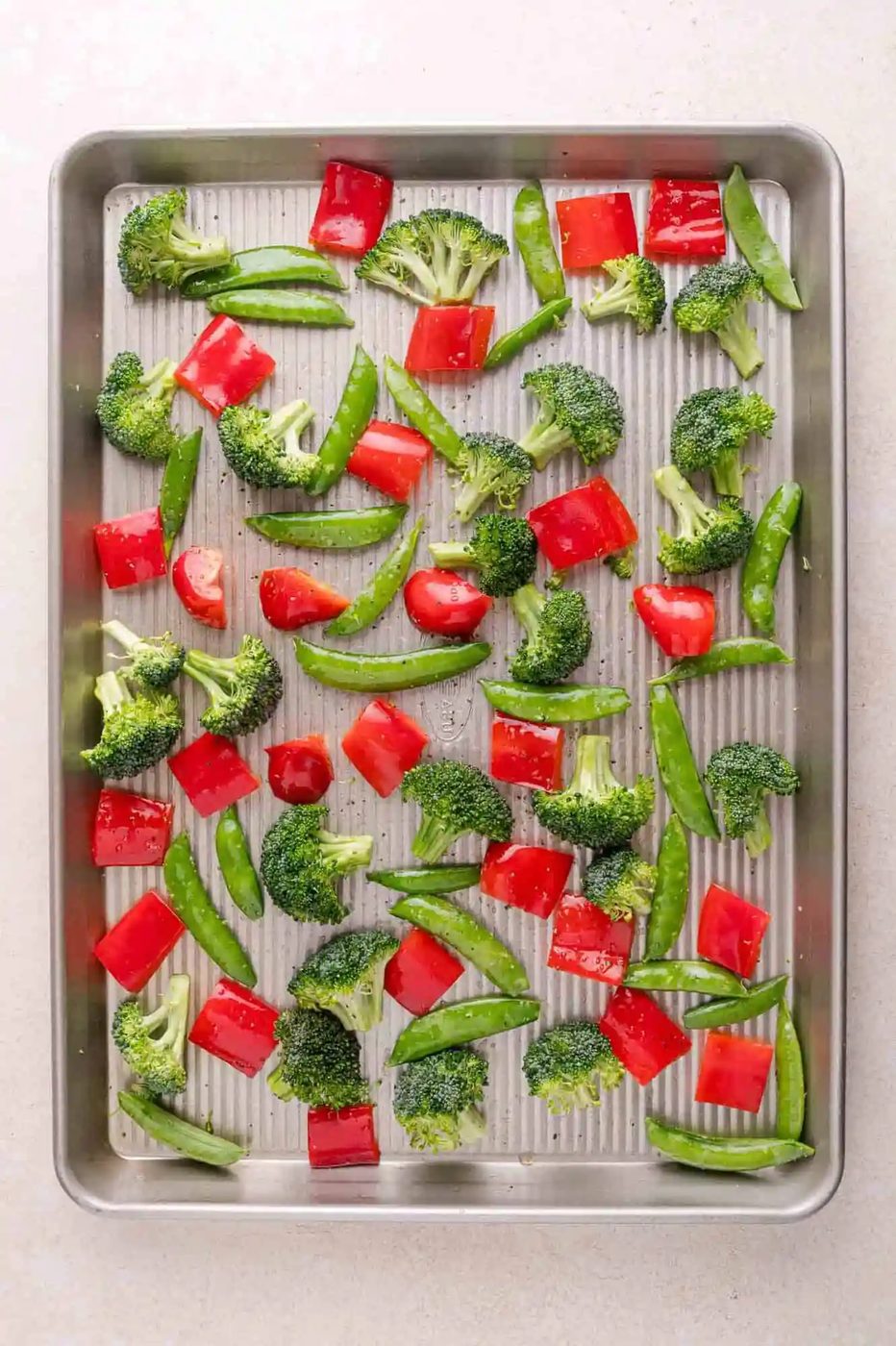 A baking sheet with raw broccoli florets, red bell pepper pieces, and snap peas spread evenly on a light-colored surface.