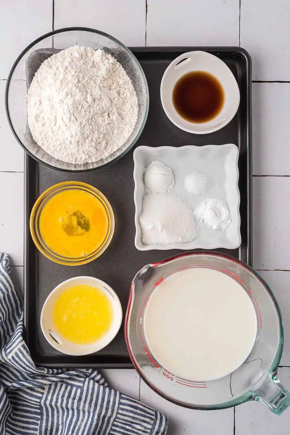 Ingredients for baking arranged on a tray: flour, vanilla extract, eggs, sugar, baking powder, salt, melted butter, and milk, with a striped cloth nearby on a tiled surface.