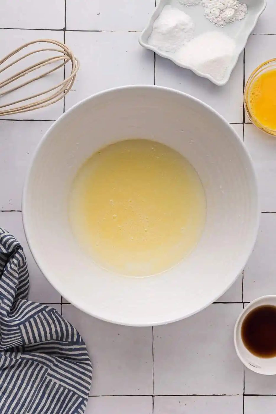 A mixing bowl with liquid ingredients surrounded by a whisk, striped cloth, small dishes of flour, sugar, egg yolks, and vanilla extract on a tiled surface.