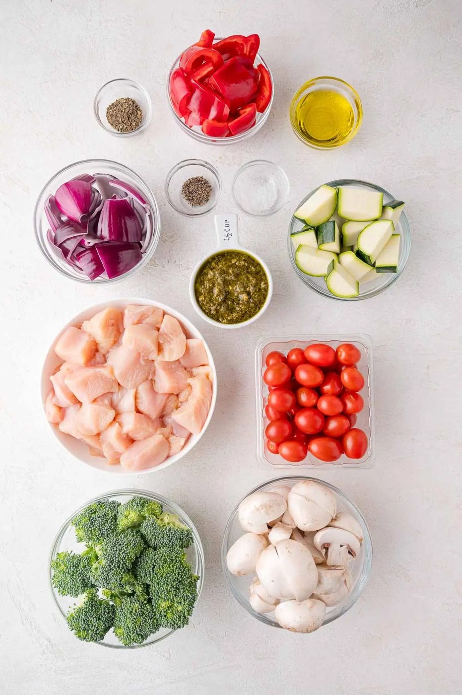 Assorted bowls of raw chicken, broccoli, mushrooms, grape tomatoes, zucchini, red onion, red bell pepper, pesto, olive oil, salt, and pepper arranged on a white surface.