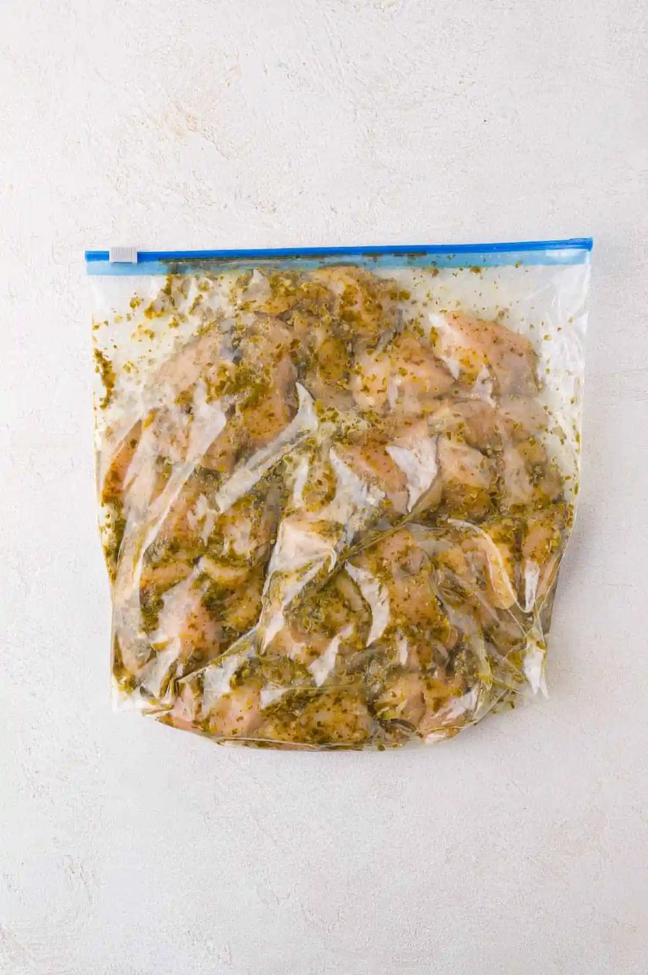Raw chicken pieces marinating in a clear resealable plastic bag with herbs and spices, placed on a light-colored surface.