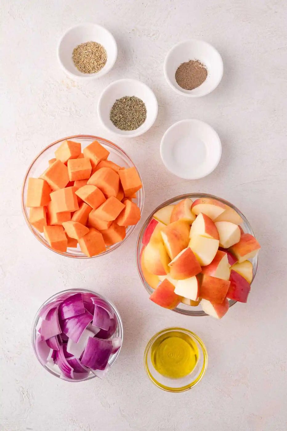 Bowls of diced sweet potatoes, chopped apples, red onion, olive oil, and small dishes of herbs and seasonings arranged on a light surface.
