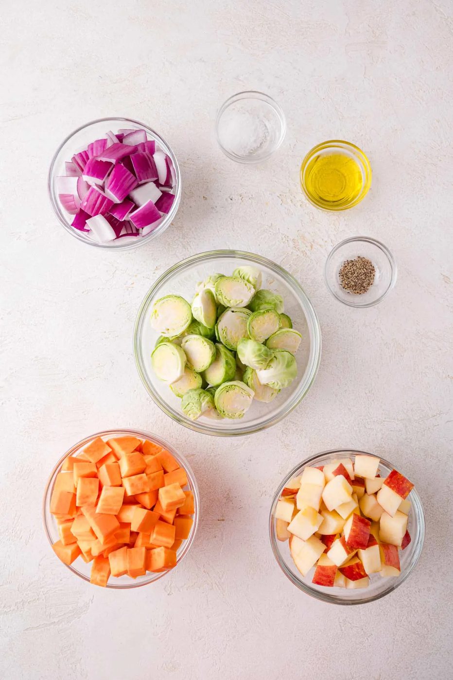 Five glass bowls with chopped red onion, halved Brussels sprouts, diced sweet potato, and diced apples, plus small bowls of oil, salt, and pepper on a light surface.