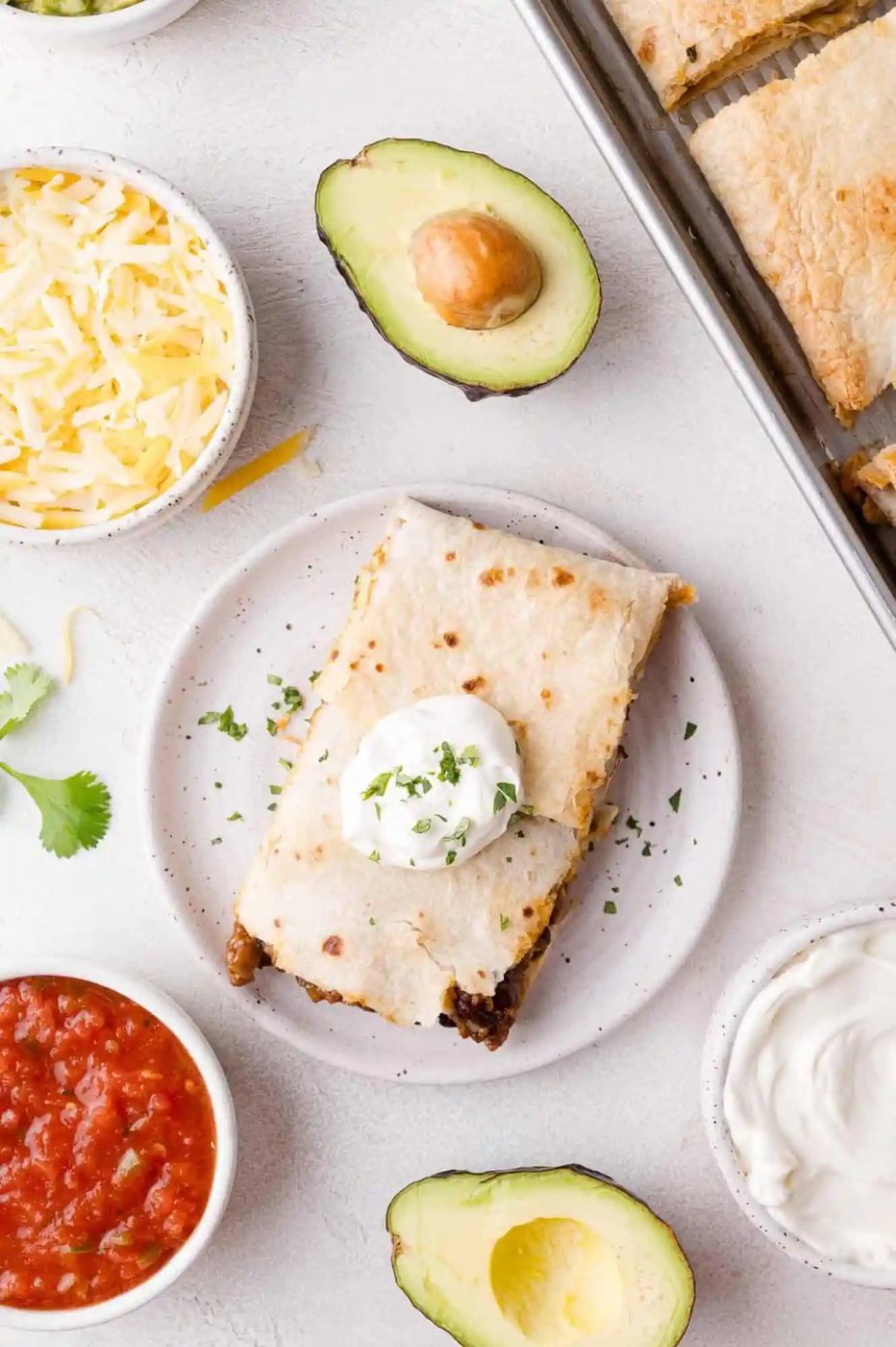 A plated sheet pan quesadilla topped with sour cream and herbs, surrounded by bowls of shredded cheese, salsa, sour cream, and avocado halves on a light surface.