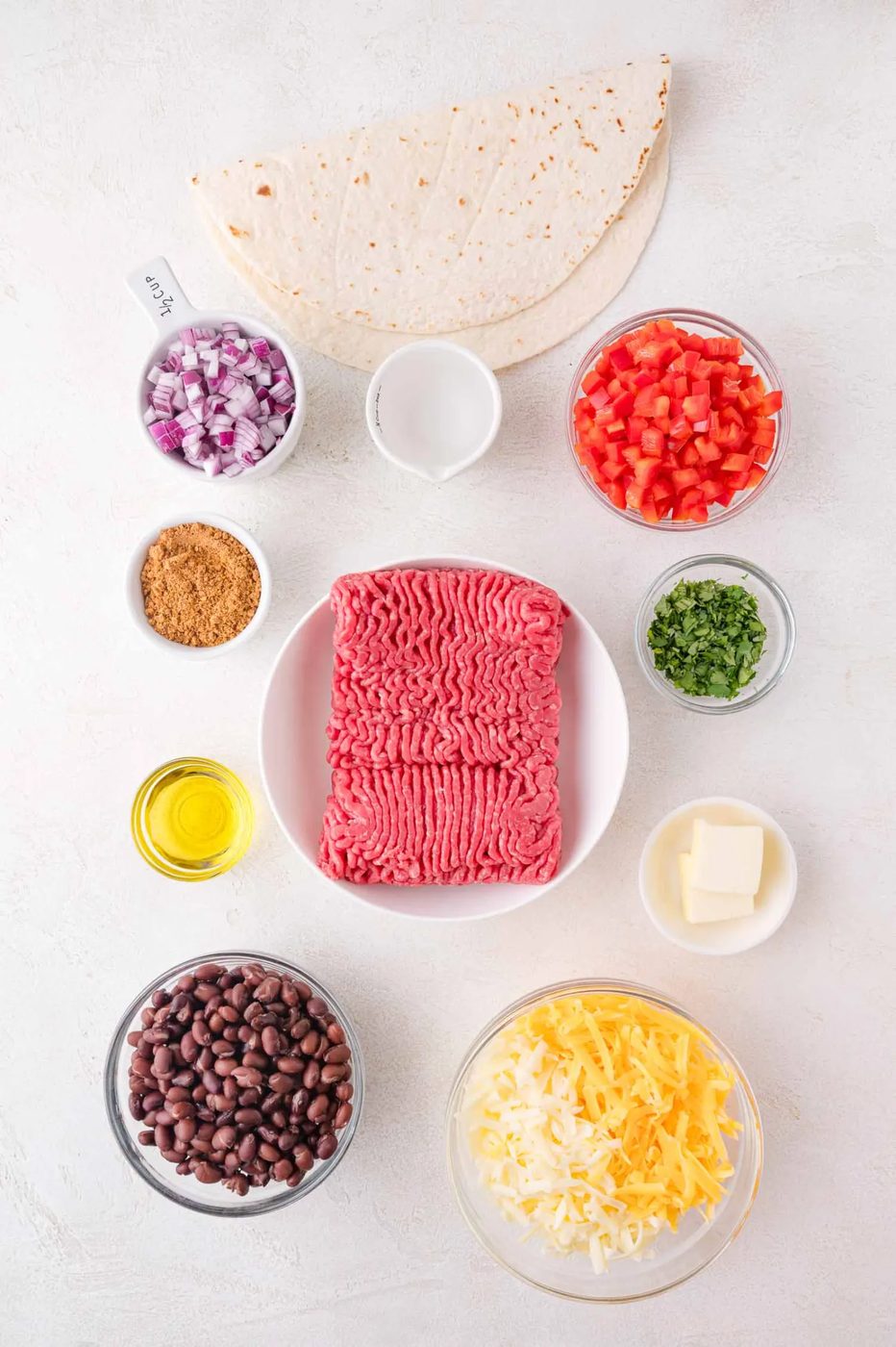 Ingredients for sheet pan quesadillas displayed: ground beef, tortillas, diced tomatoes, black beans, shredded cheese, chopped cilantro, diced onions, olive oil, sour cream, taco seasoning, and butter.
