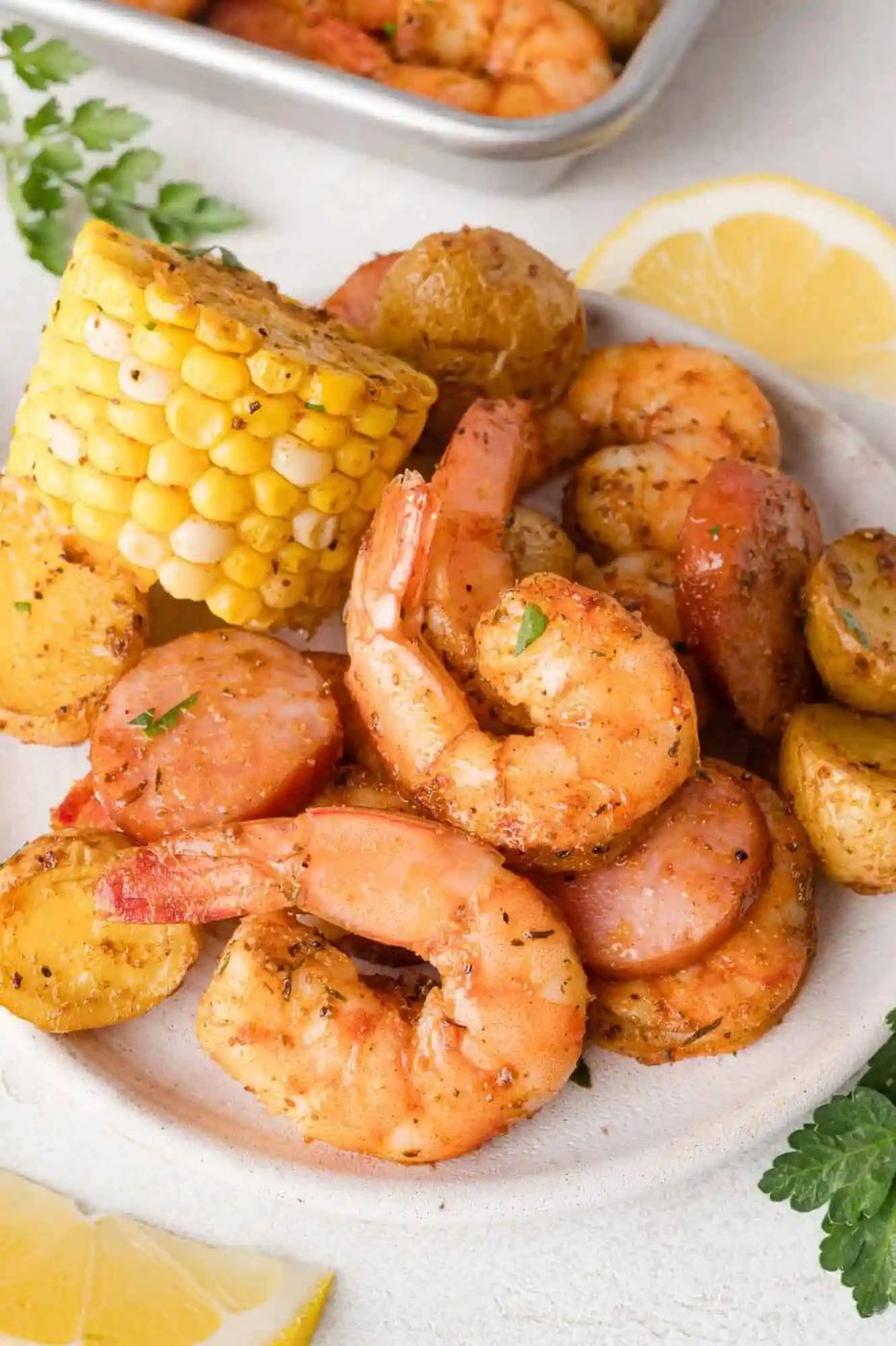 Plate of cooked shrimp, sausage slices, corn on the cob, and seasoned potatoes, garnished with herbs, with lemon slices and parsley on the side.