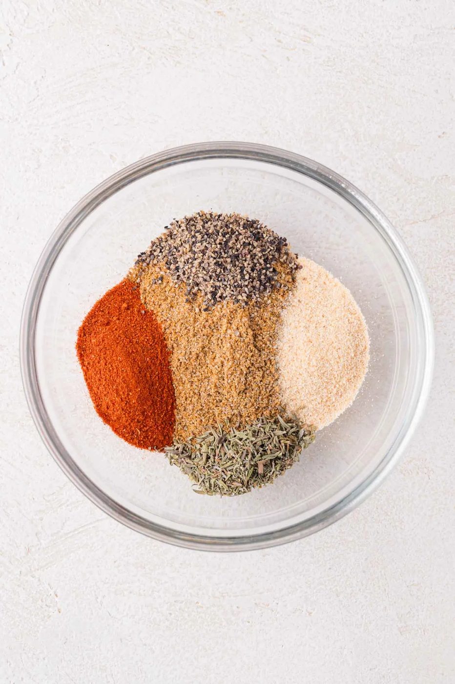 A glass bowl containing separate piles of dried spices, including paprika, black pepper, garlic powder, thyme, and brownish seasonings, on a light surface.