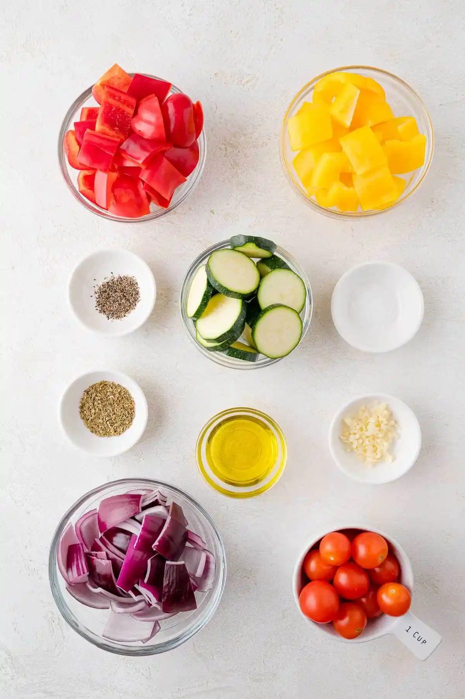 Bowls with diced red and yellow bell peppers, sliced zucchini, cherry tomatoes, chopped red onion, olive oil, minced garlic, and small bowls of salt and pepper on a white surface.