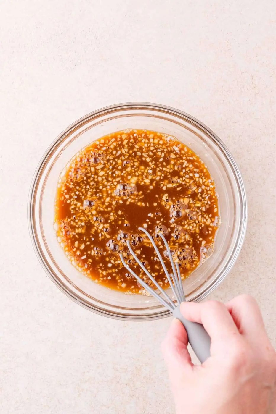 A hand holding a whisk mixes a brown sauce with visible garlic in a clear glass bowl on a light-colored surface.