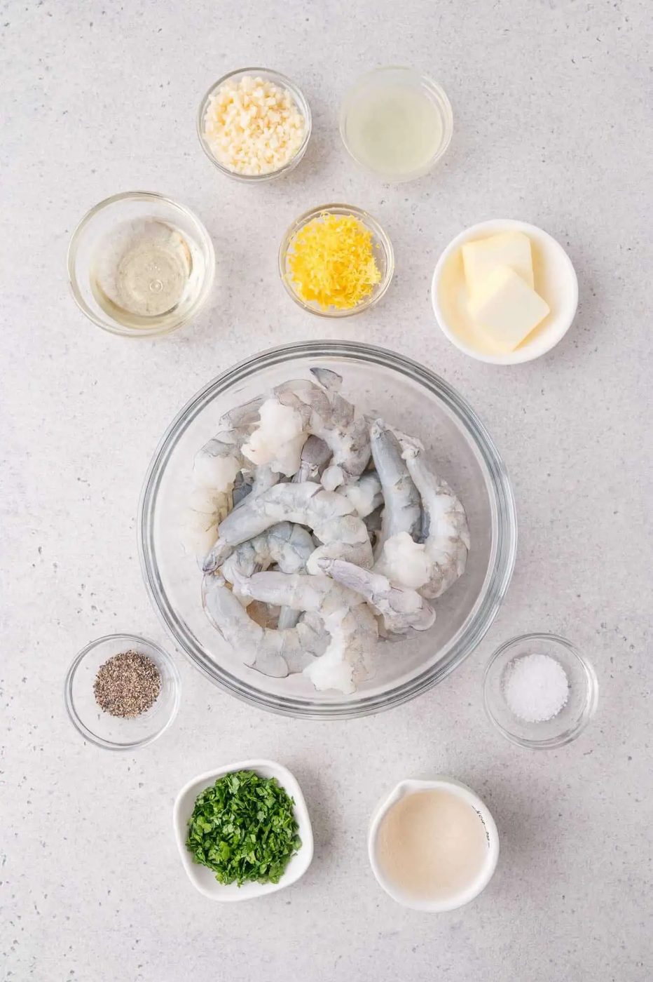 Raw shrimp in a glass bowl surrounded by bowls of chopped garlic, lemon zest, lemon juice, butter, salt, pepper, parsley, white wine, and cream on a light countertop.