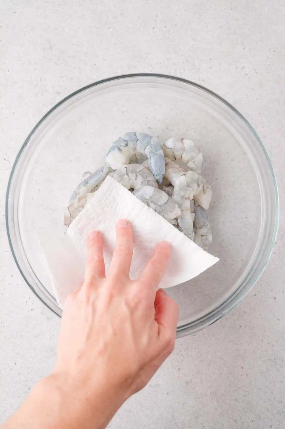 A hand uses a paper towel to pat dry raw, peeled shrimp in a clear glass bowl on a light-colored surface.