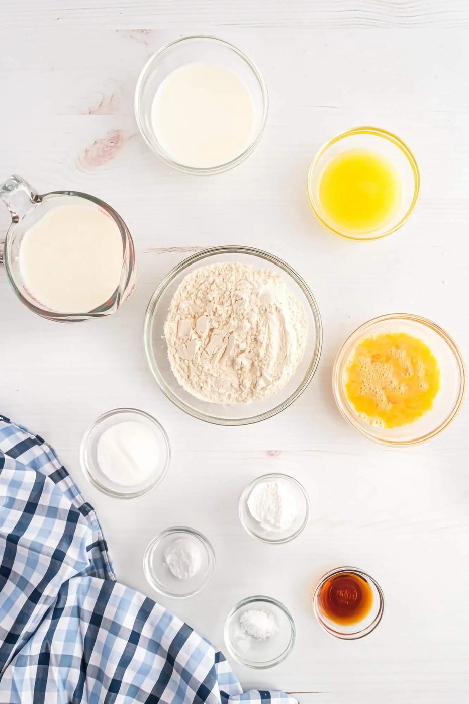 Various bowls containing flour, beaten eggs, melted butter, milk, vanilla extract, salt, baking powder, and sugar on a white surface with a checkered cloth in the corner.