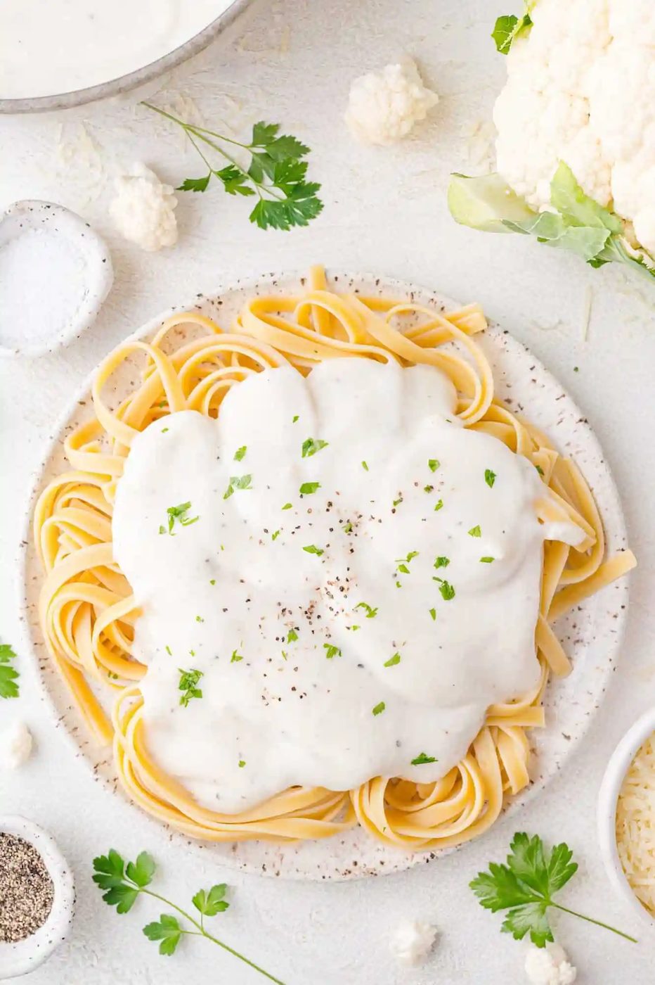 A plate of fettuccine pasta topped with creamy white sauce, garnished with black pepper and parsley. Surrounding the plate are small bowls and parsley leaves on a light surface.