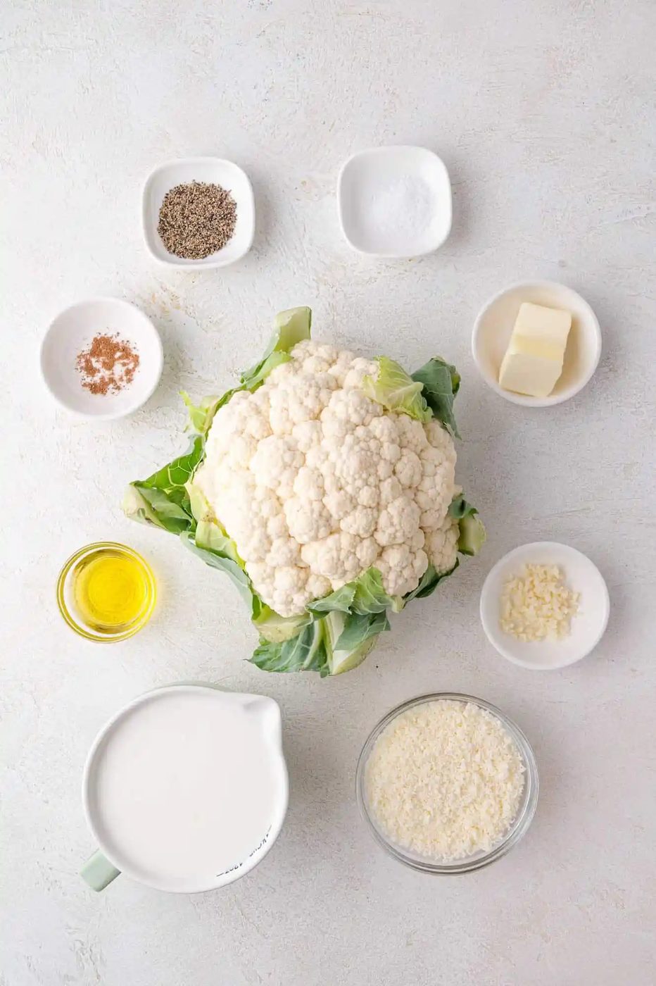 Top-down view of cauliflower surrounded by bowls containing milk, olive oil, butter, grated cheese, minced garlic, and spices on a white surface.