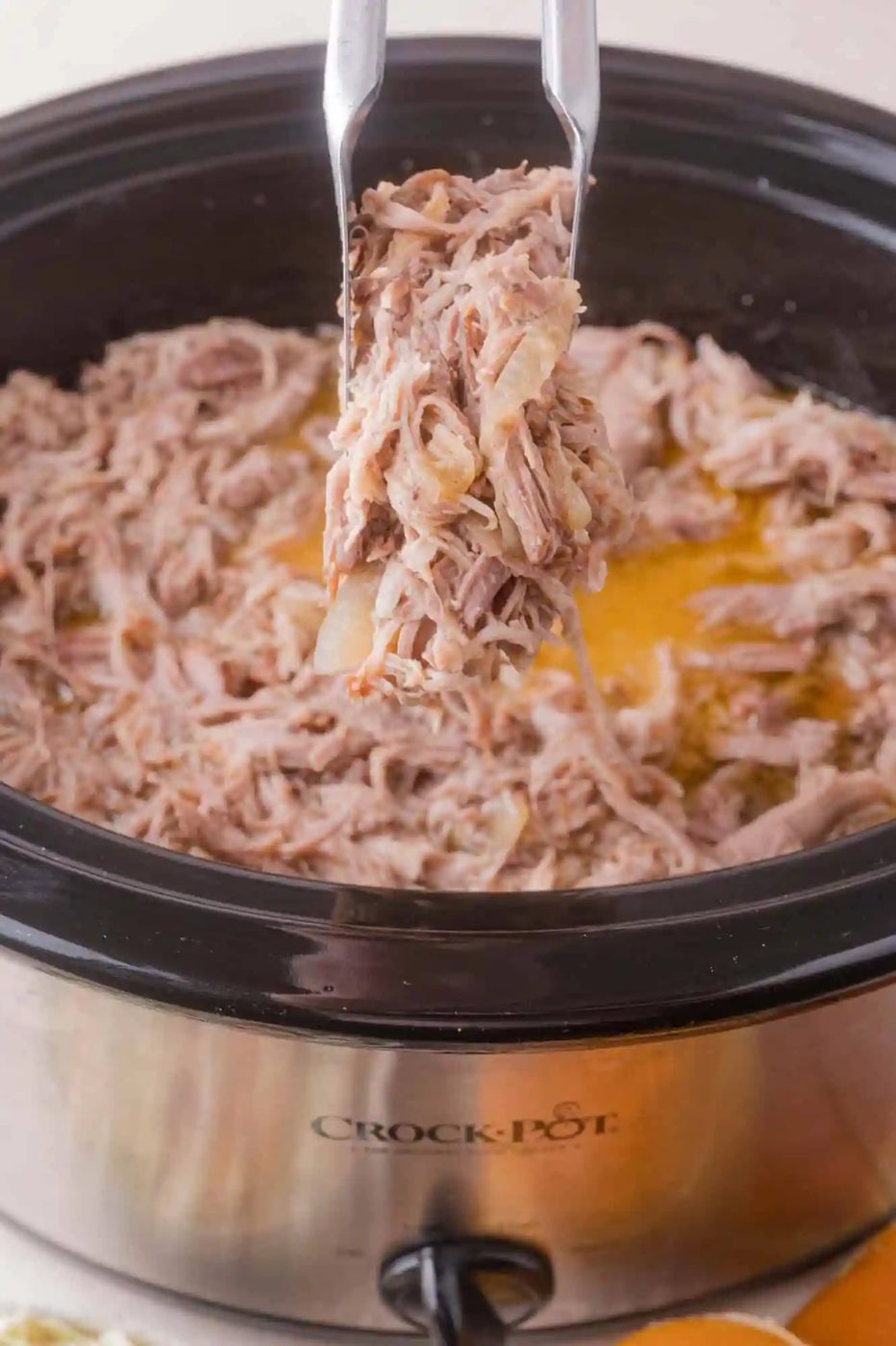 Shredded pork being lifted with tongs from a slow cooker filled with cooked meat and juices.