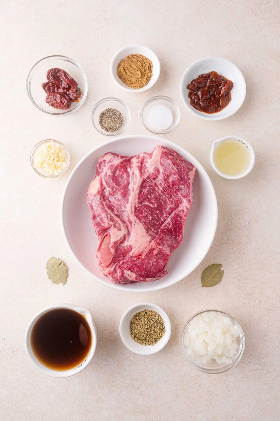 A raw marbled cut of beef in a bowl surrounded by small bowls containing various spices, chopped onion, sauce, and other ingredients on a light surface.