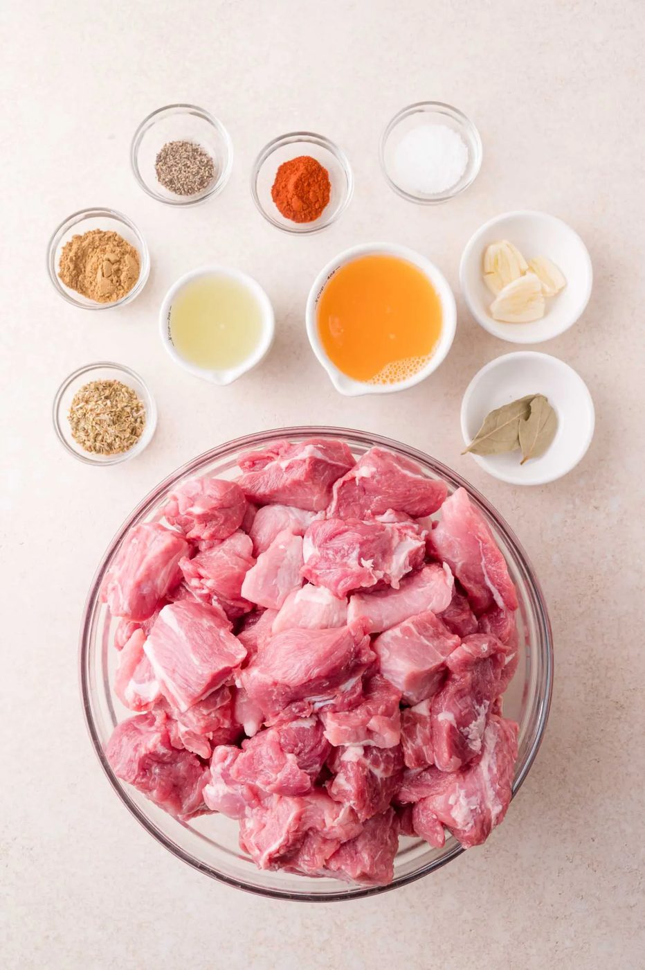 A glass bowl filled with raw pork chunks is surrounded by small bowls of spices, herbs, garlic cloves, vinegar, salt, pepper, and bay leaves on a light surface.