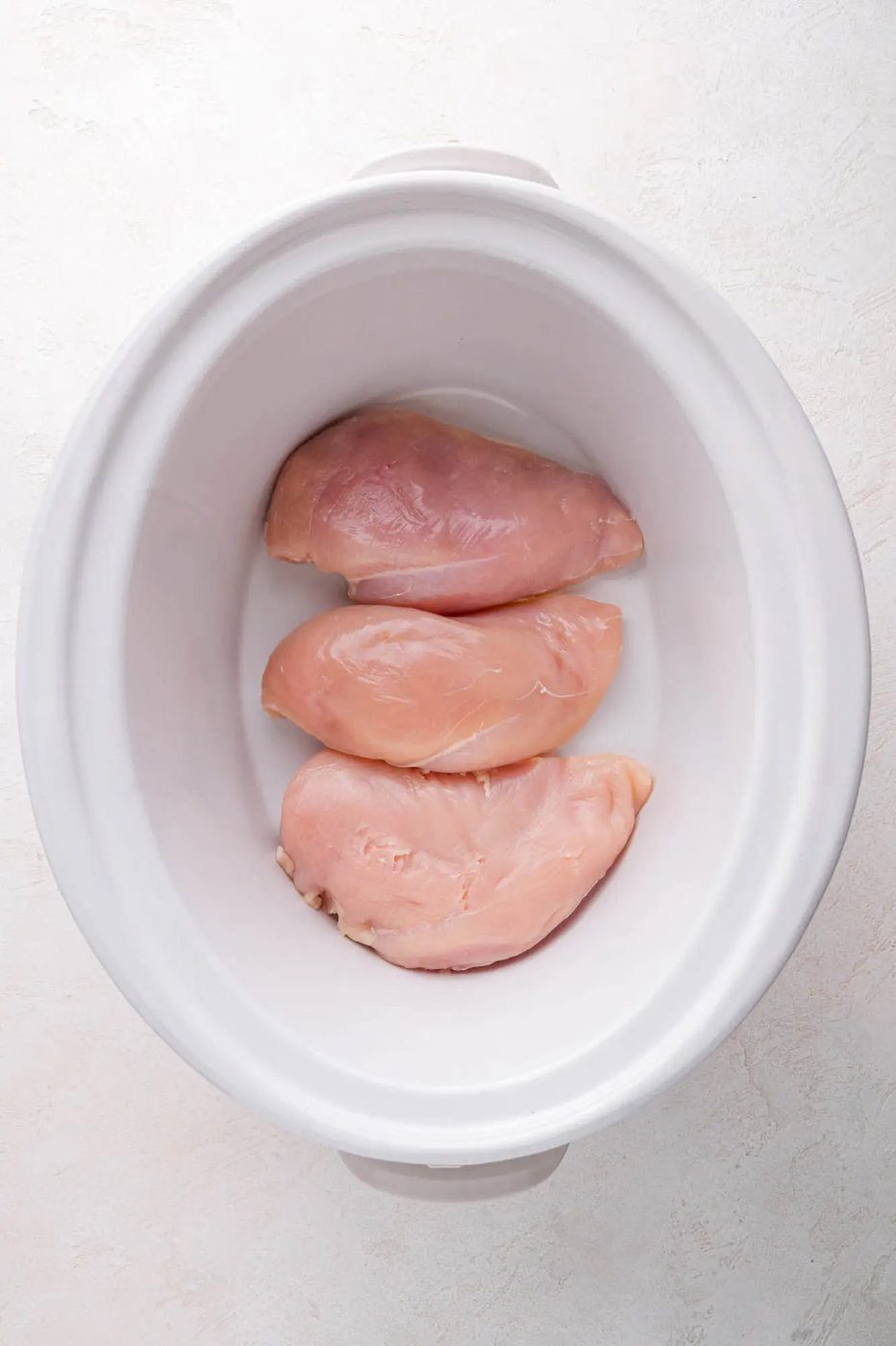 Three raw chicken breasts placed inside a white slow cooker on a light-colored surface.