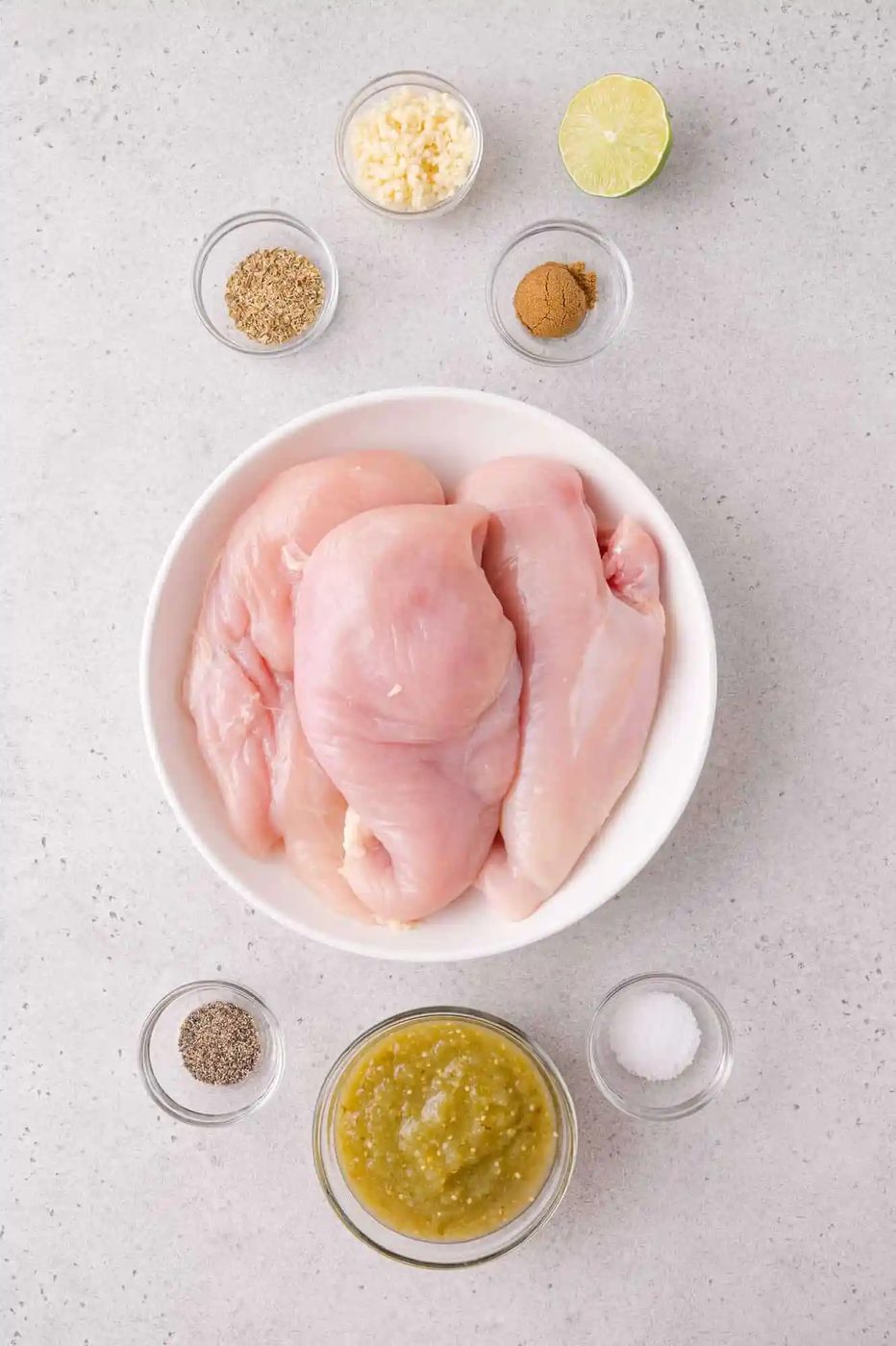 Raw chicken breasts in a white bowl surrounded by small bowls of seasonings, minced garlic, salsa verde, half a lime, pepper, and salt on a light countertop.