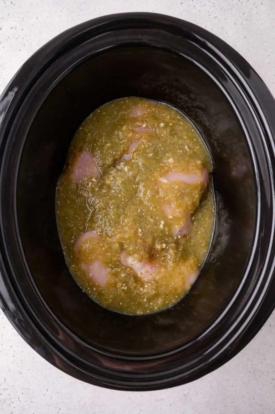 Raw chicken breasts covered in salsa verde inside a black slow cooker, photographed from above on a light-colored surface.