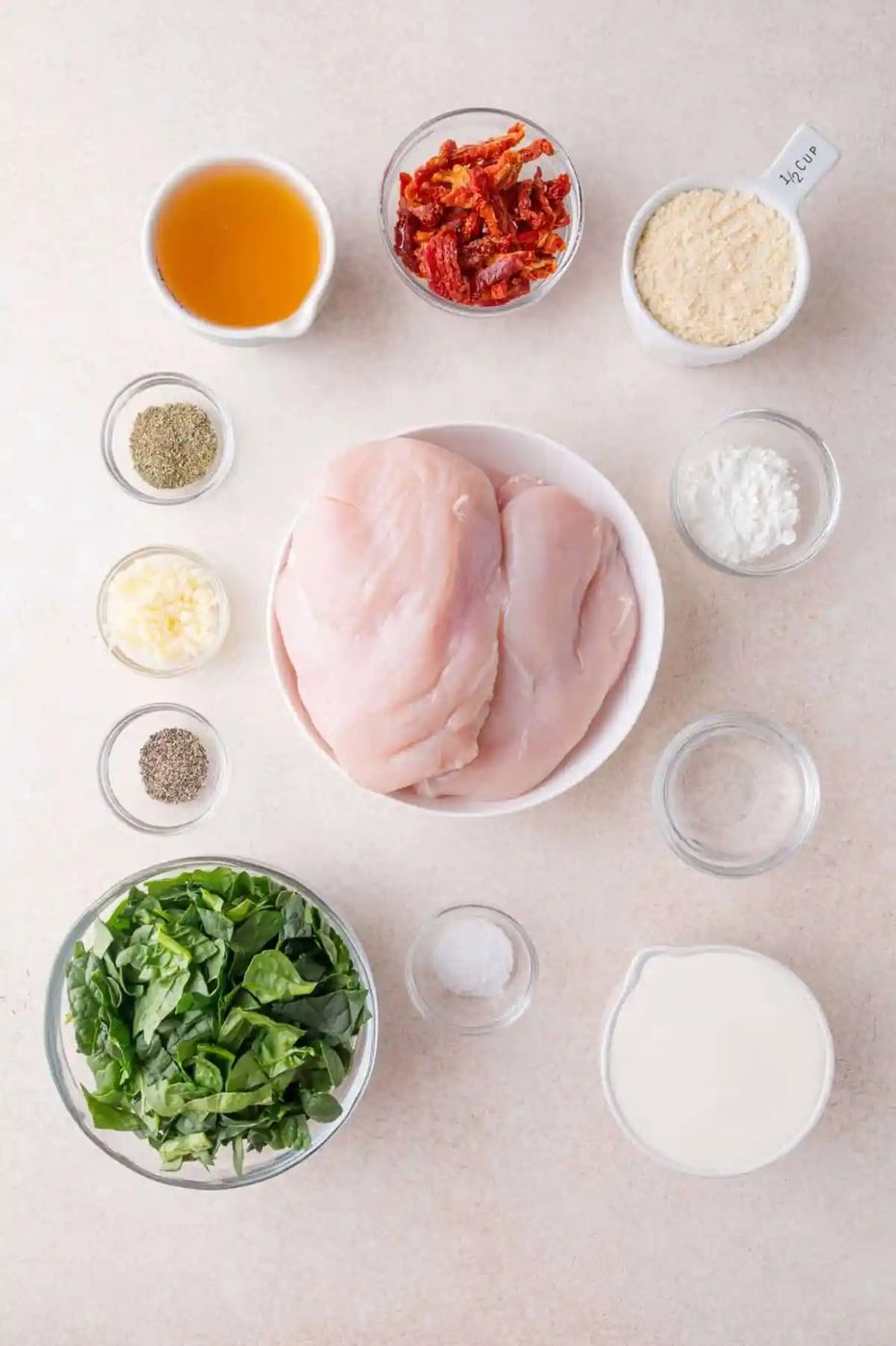 Top-down view of chicken breasts, fresh spinach, cream, sun-dried tomatoes, parmesan cheese, garlic, salt, pepper, Italian seasoning, and cornstarch in individual bowls on a countertop.