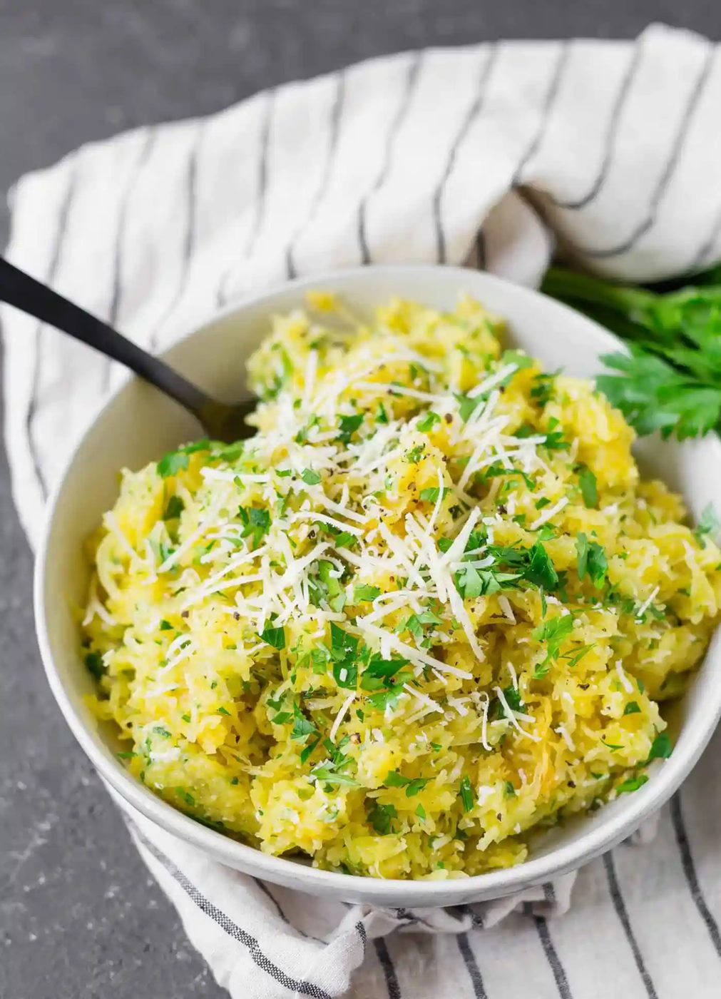 A bowl of spaghetti squash topped with grated cheese and chopped parsley, with a fork and a striped cloth underneath.