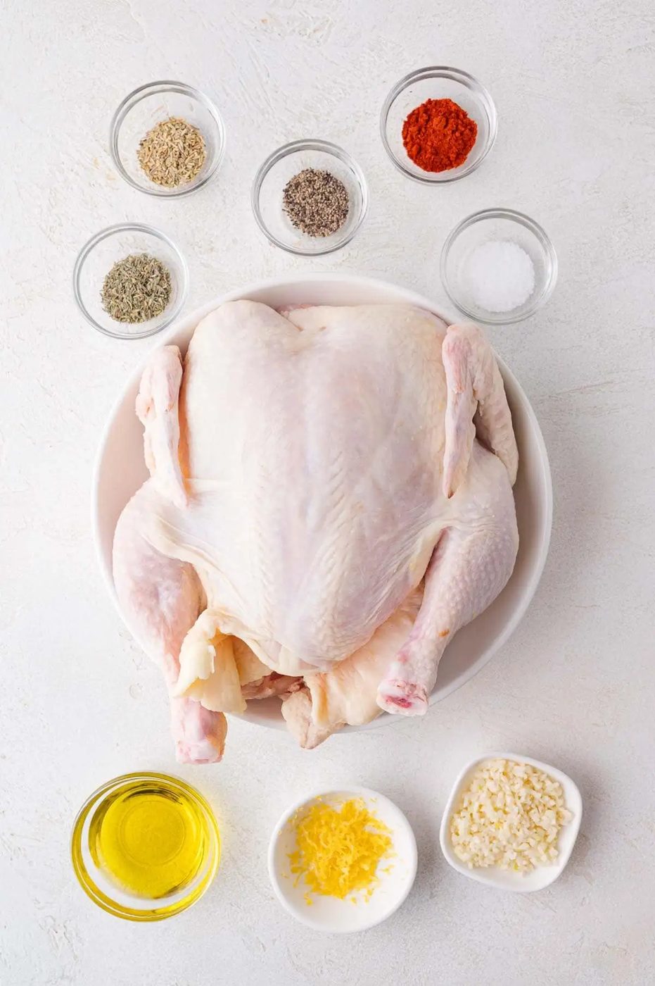 Whole raw chicken in a bowl surrounded by spices, oil, garlic, lemon zest, and salt on a white surface.