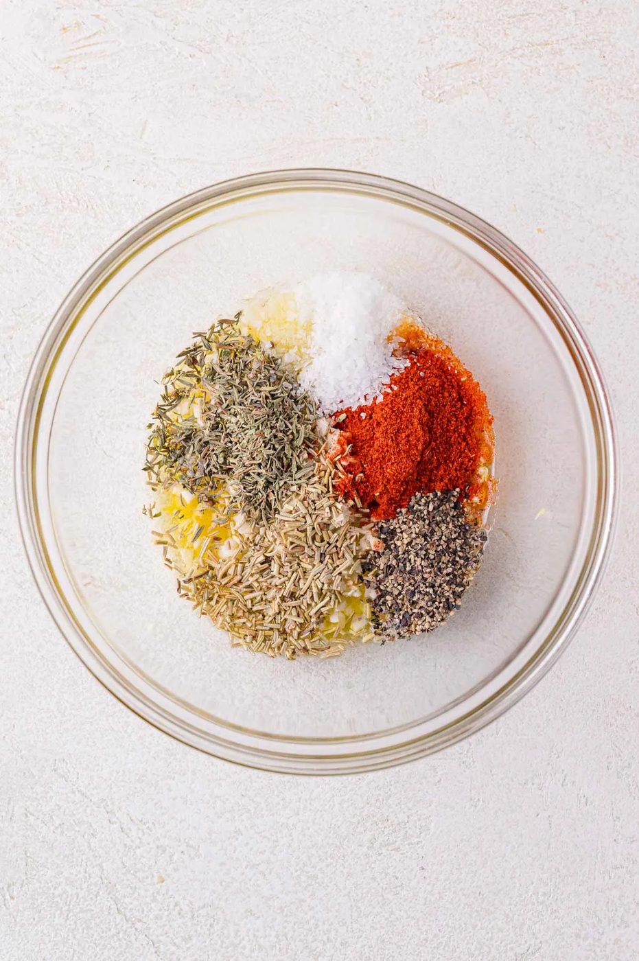 A glass bowl containing dried herbs, black pepper, paprika, salt, and olive oil on a light background.