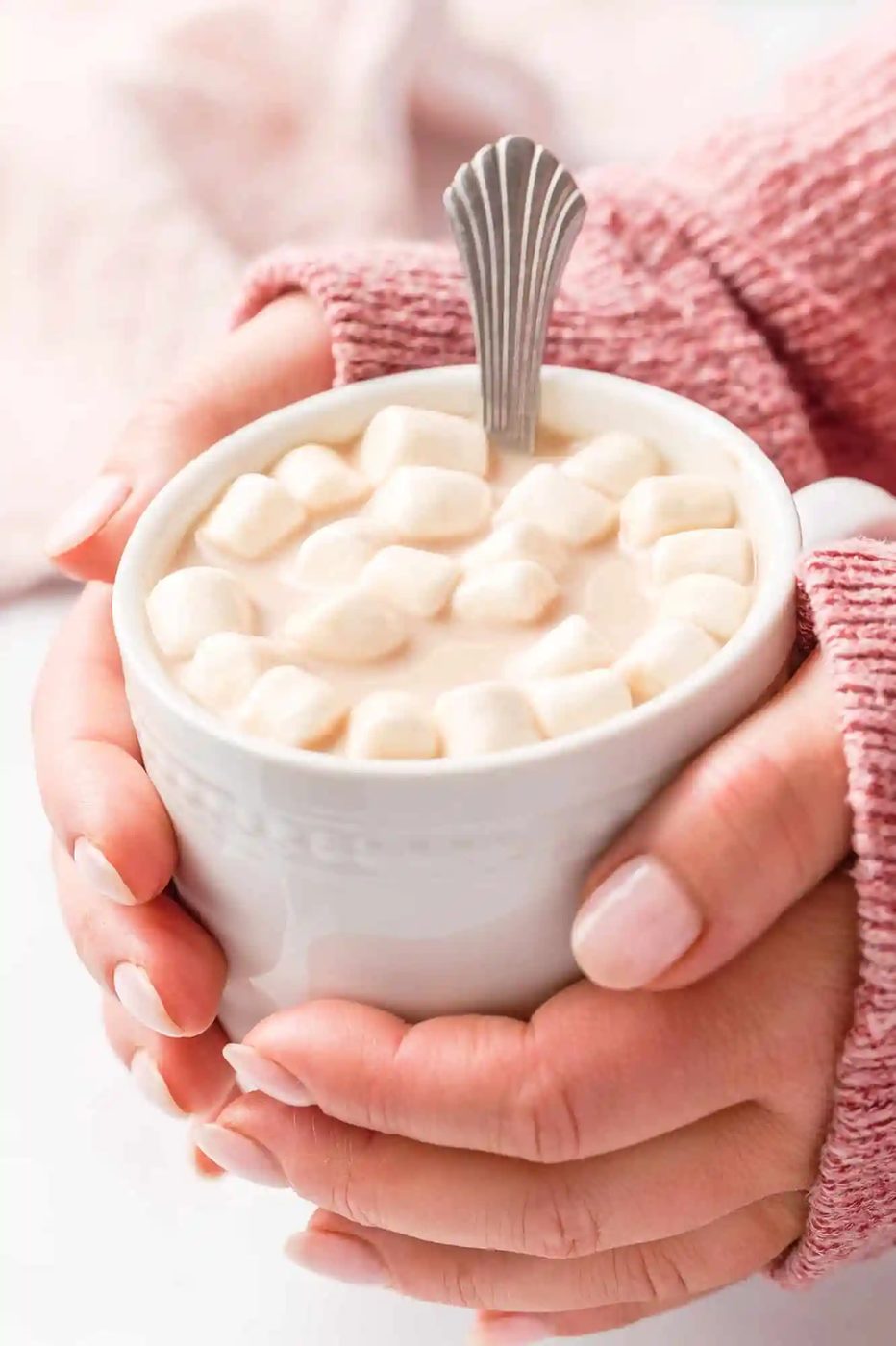 Hands holding a white mug filled with hot chocolate topped with mini marshmallows and a spoon, wearing a pink knitted sweater.