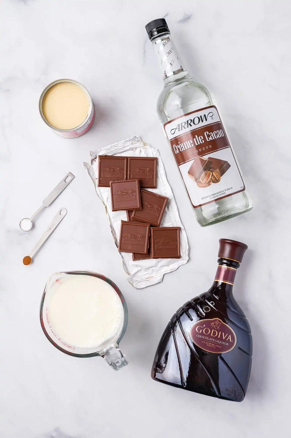 Ingredients laid out on a surface include a bottle of Crème de Cacao, a can of condensed milk, chocolate squares, a measuring cup of cream, a bottle of Godiva liqueur, and measuring spoons.