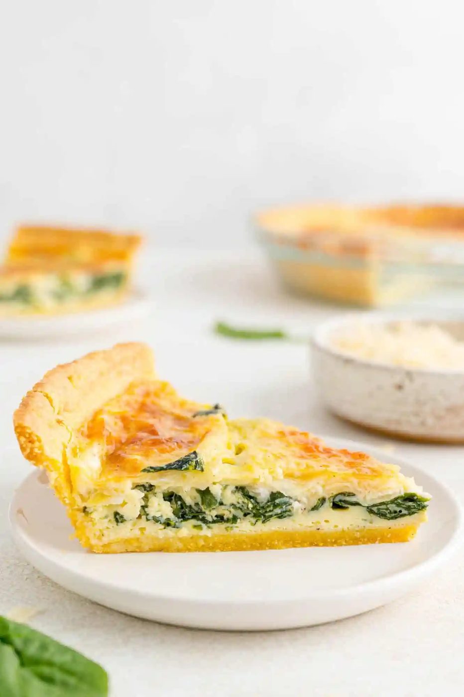 Slice of quiche on a white plate, more quiche in background.