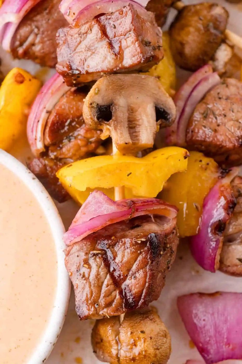 Close-up of a skewer with grilled beef, yellow bell pepper, red onion, and mushroom next to a bowl of dipping sauce.