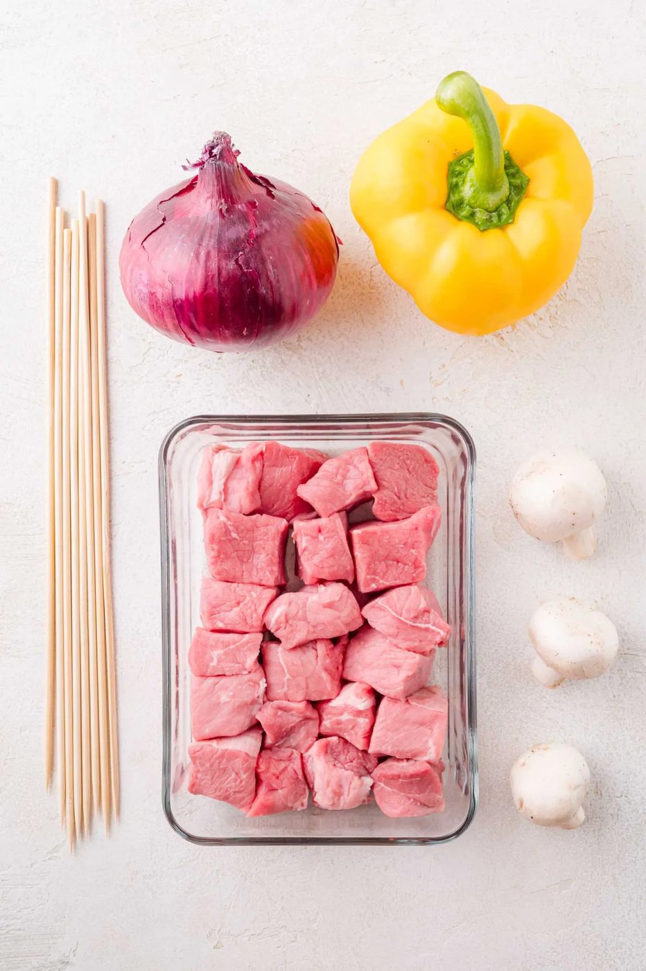 Glass container with raw steak cubes, a red onion, yellow bell pepper, two white mushrooms, and wooden skewers arranged on a white surface.