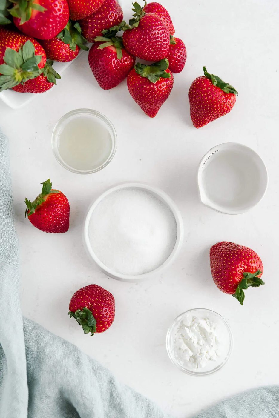 A flat lay of fresh strawberries, a bowl of sugar, small jars of lemon juice and water, and a small bowl of cornstarch on a white surface.