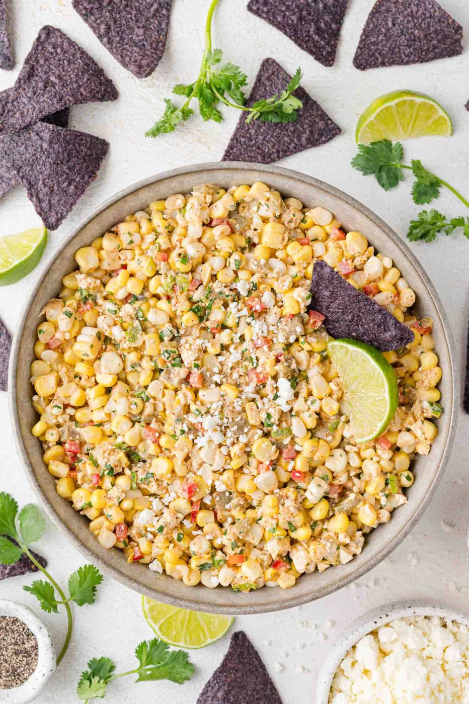 Bowl of Mexican street corn dip garnished with lime slices and crumbled cheese, surrounded by blue tortilla chips and fresh cilantro on a white surface.