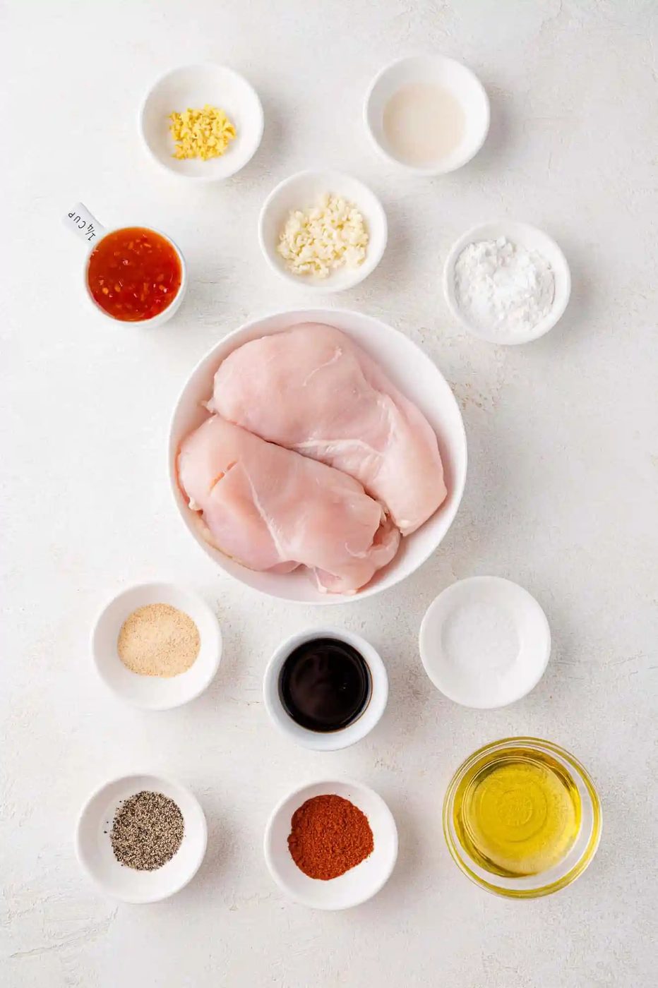 Raw chicken breasts surrounded by small bowls of ingredients including garlic, chili sauce, soy sauce, oil, and spices.