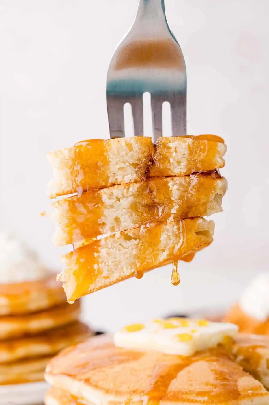 A fork holding a stack of syrup-drizzled pancakes, with a stack of pancakes topped with butter visible in the background.
