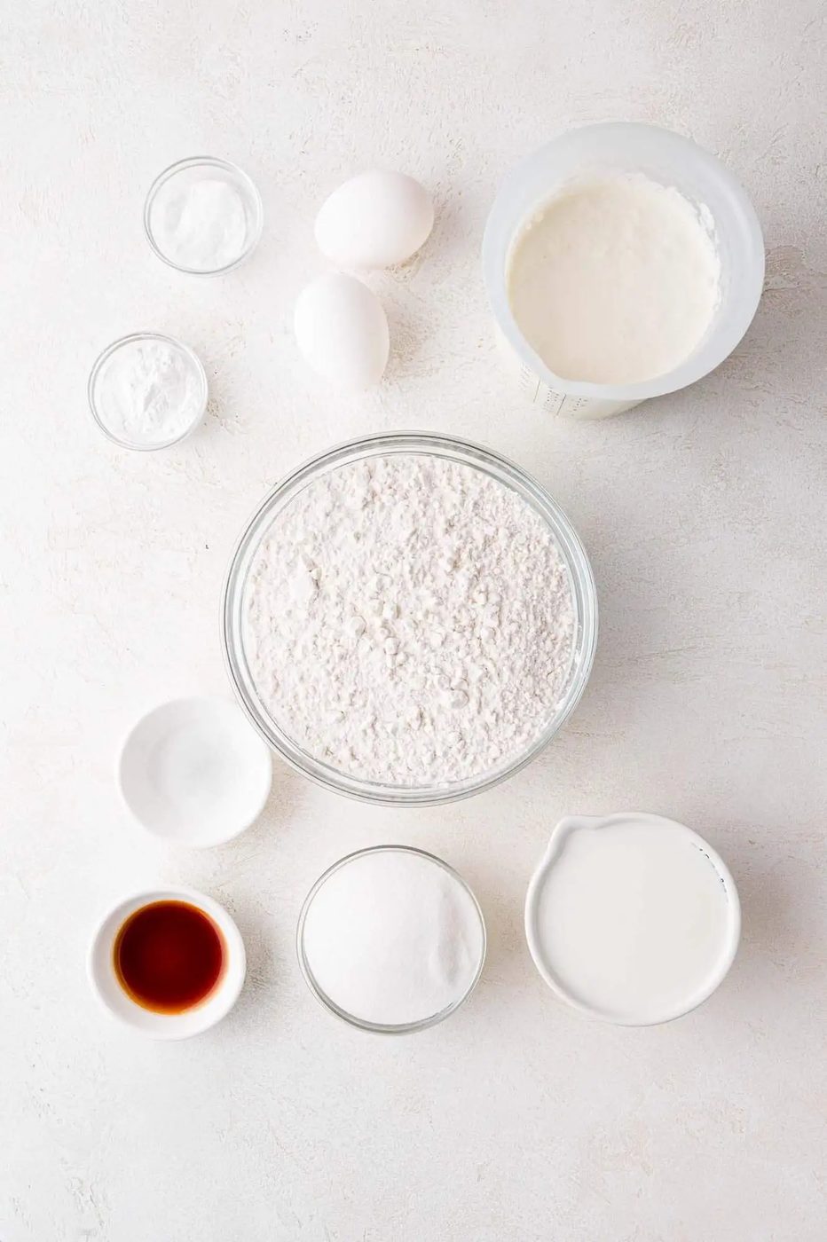 Baking ingredients on a table: flour, sugar, milk, vanilla extract, eggs, and bowls of baking soda, powder, and cream.