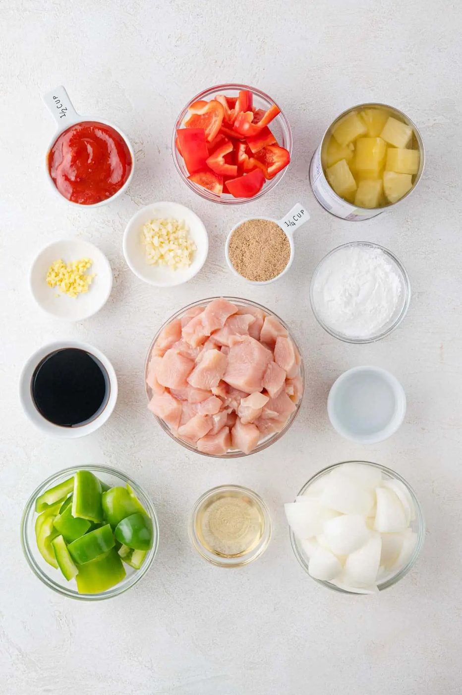Ingredients for a chicken dish arranged on a surface, including diced chicken, bell peppers, onions, garlic, soy sauce, tomato, pineapple, ginger, and various seasonings in small bowls.
