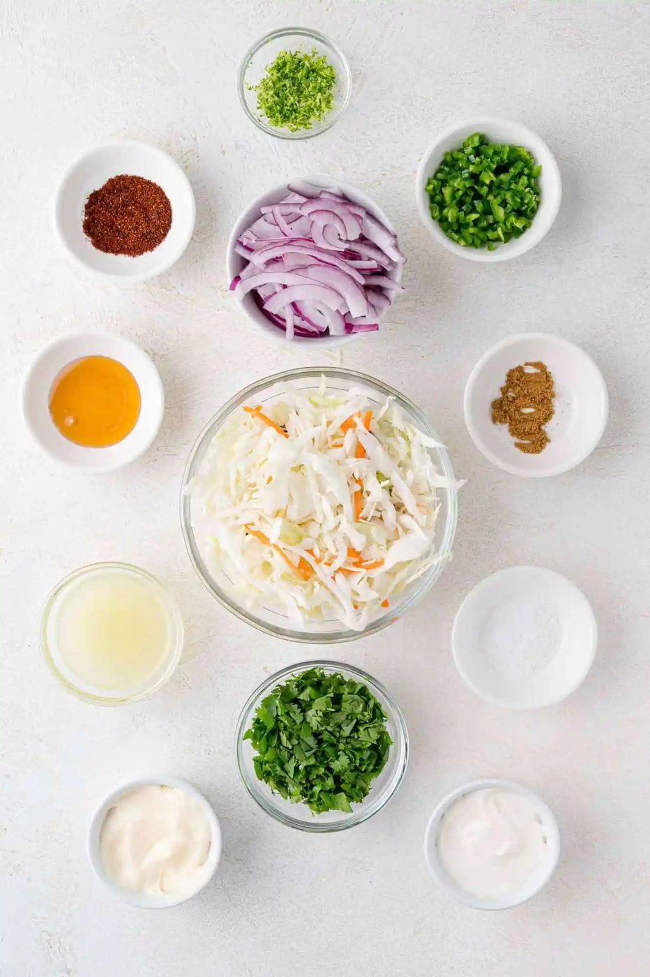 A variety of ingredients for taco slaw arranged in small bowls on a white surface, including cabbage, carrots, red onion, herbs, spices, mayonnaise, lime juice, and sour cream.