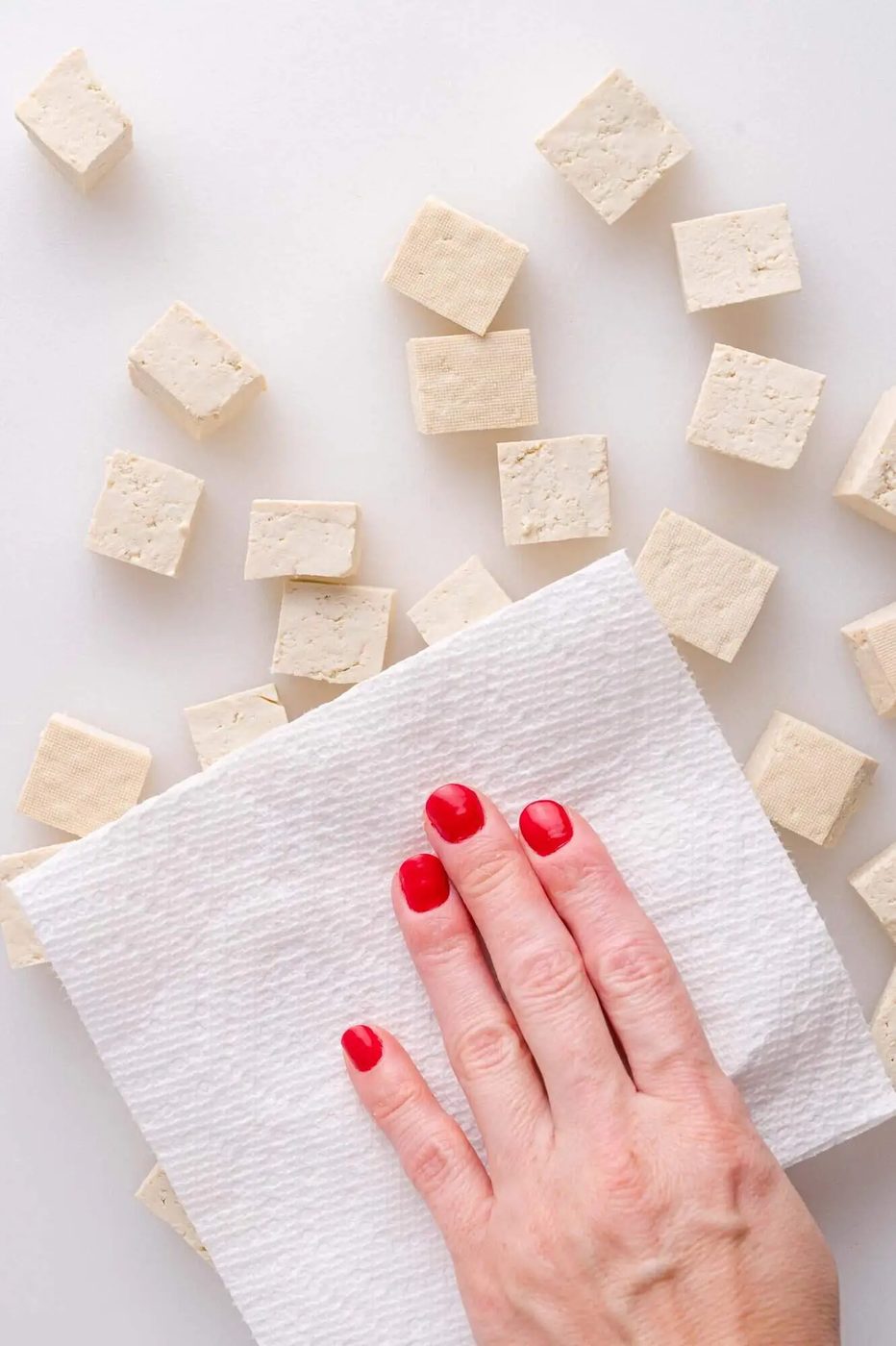 A hand with red nail polish presses a paper towel onto cubes of tofu on a white surface.