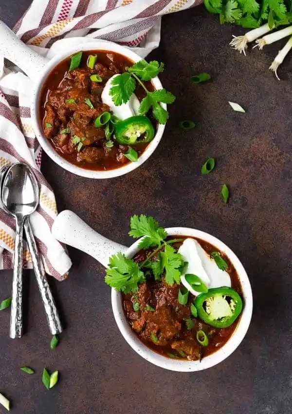 Overhead of chili in round white bowl with handle, topped with cilantro chopped scallions, a slice of pepper, and sour cream.
