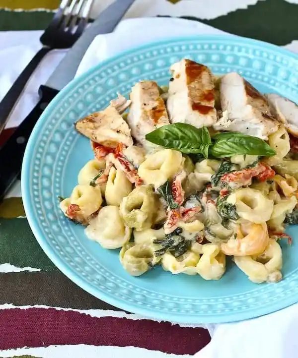 Shallow blue bowl with tortellini and sun-dried tomato sauce, garnished with fresh basil and served with sliced cooked chicken