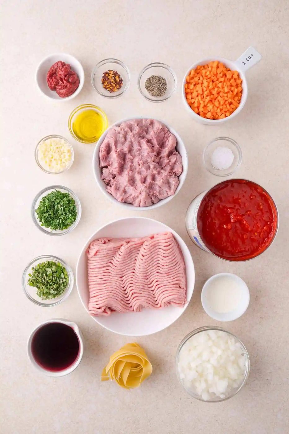 Ingredients for a pasta dish arranged on a countertop, including ground meat, tomato sauce, vegetables, herbs, spices, olive oil, cream, and lasagna noodles in separate bowls.