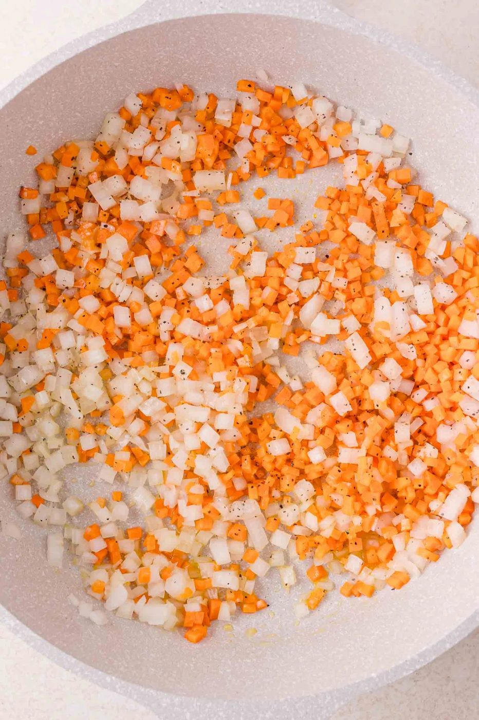 Diced carrots and onions being sautéed in a pan with visible black pepper seasoning.